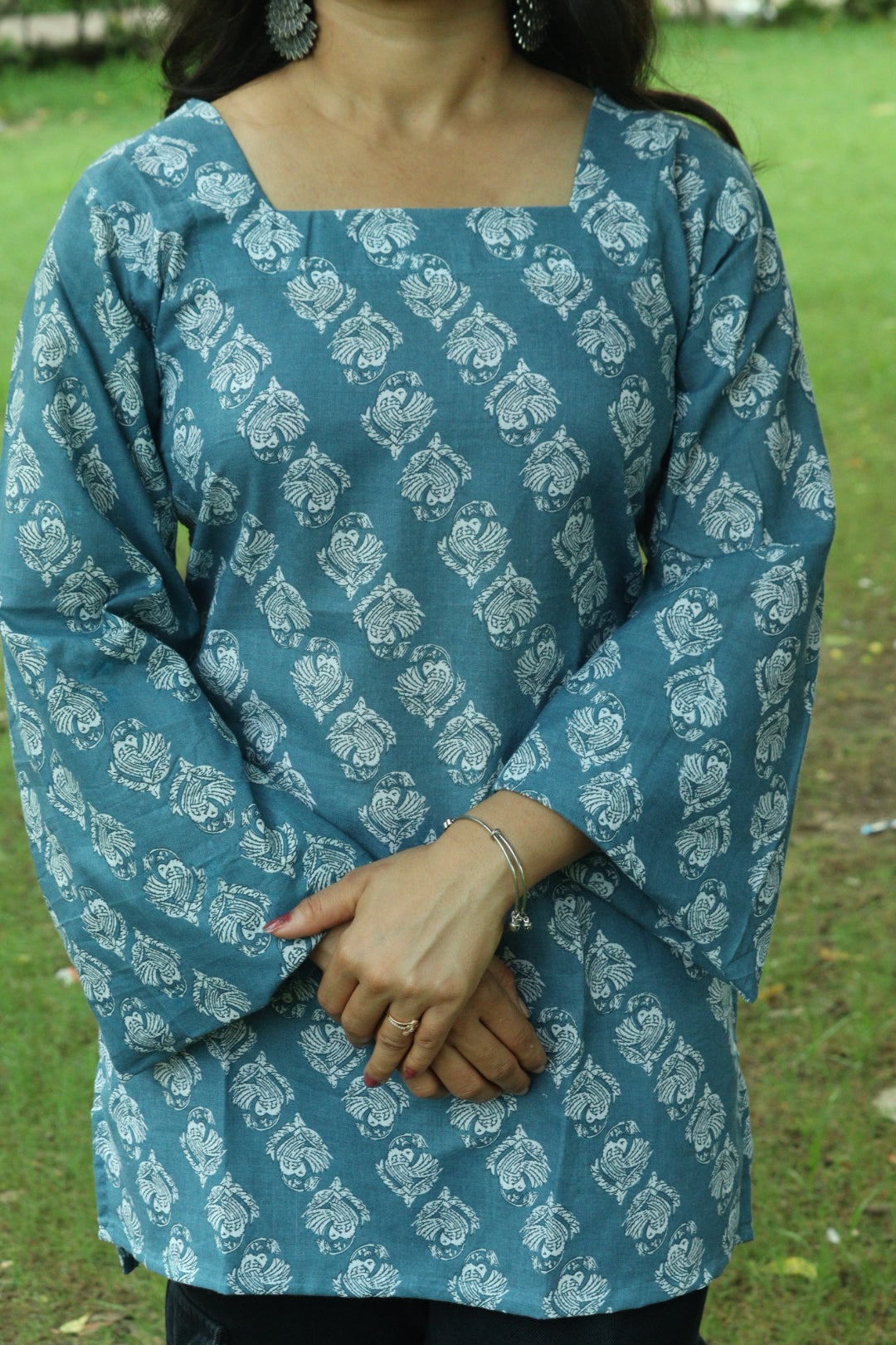 A woman wearing a teal blue block-printed kurti with a square neckline, photographed outdoors against soft greenery.