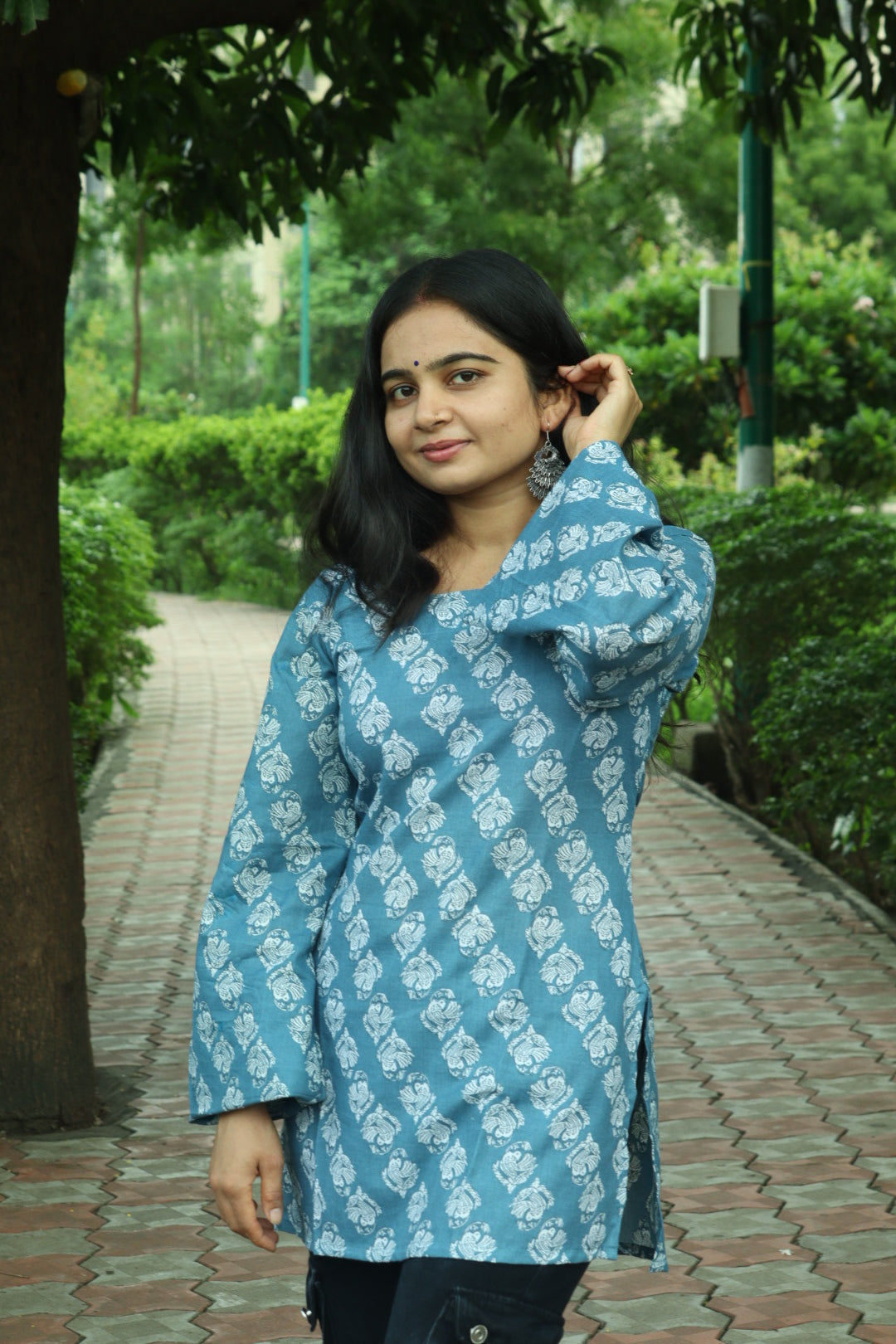 A woman wearing a light blue and white patterned kurta standing outdoors on a paved path with green trees in the background.