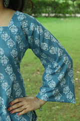 A woman wearing a teal blue short kurti with ivory block prints and side slits, standing on a paved garden path.